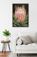 Framed canvas artwork of a pink protea flower with green leaves on a dark background, hanging above a white sofa and next to a small plant on a wooden table.