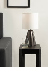 Modern scalloped base table lamp in dark bronze with a white cylindrical fabric shade, placed on a black side table next to a dark sofa arm.