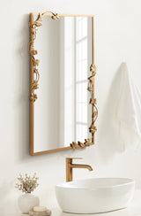 Rectangular mirror with a gold metal frame featuring floral and vine accents on two edges, mounted above a white oval vessel sink and gold faucet.