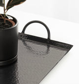 Black hammered metal rectangular tray with rounded handles on each short side, shown with a black planter holding a green plant on a white surface.