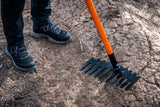 Person wearing black boots standing on dry, cracked soil next to a black metal rake with an orange handle lying on the ground.