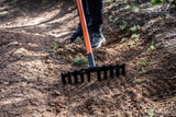 Close-up of an orange-handled rake tilling dry soil on a sunny dirt path with scattered green plants and a person wearing black pants and shoes standing behind it.