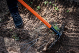 Person wearing black shoes using an orange-handled tool to dig and clear roots and soil in a garden area with dry earth and small plants.