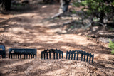 Six black combs arranged in a row on a sandy dirt path with scattered shadows and green shrubbery in the background, highlighting their varying heights and teeth shapes.