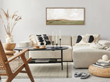 Minimalist living room with beige sectional sofa adorned with black and cream patterned pillows, wooden-framed landscape painting, woven chair, round rattan ottoman, and black oval coffee table on beige rug.