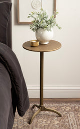 Round bronze-colored metal side table with a central pedestal and three-legged base, holding a cream ceramic vase with green leafy plant and a small gold candle holder.