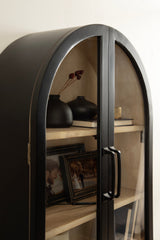 Black metal arch-shaped wall cabinet with glass doors revealing light wood shelves holding black vases and framed photos inside.