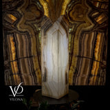 Tall, rectangular white onyx floor lamp with layered, translucent brown and gray banding, set against a dark abstract patterned wall emphasizing its glow.
