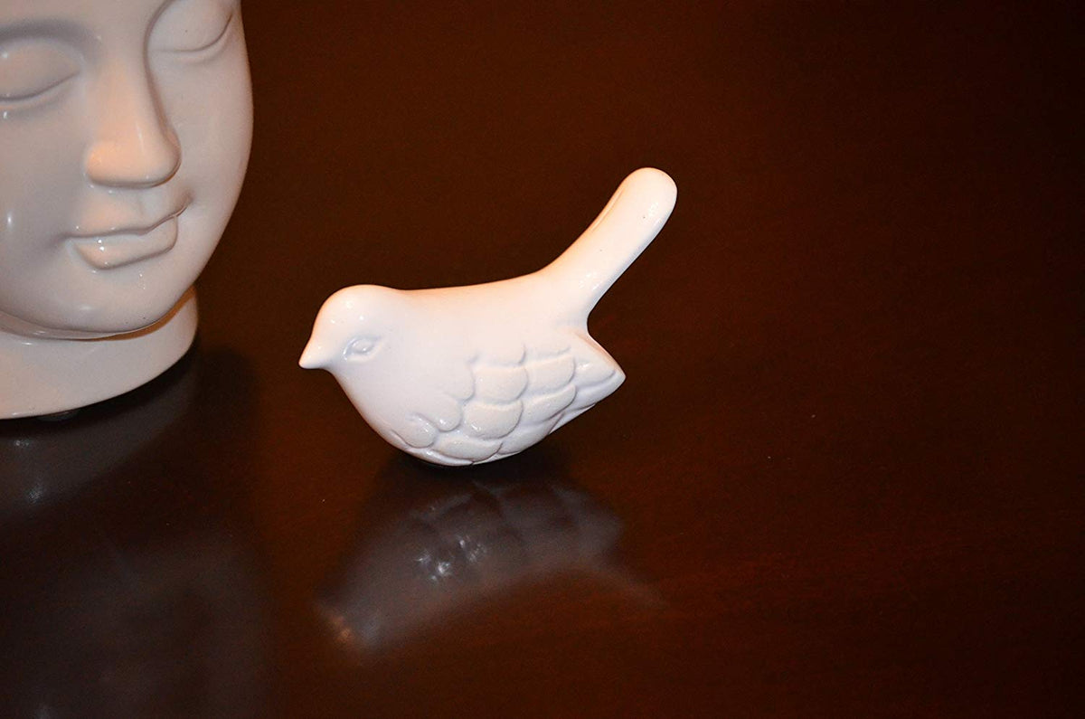 Small white ceramic bird figurine with textured wing details placed on a dark reflective surface next to part of a serene human face sculpture.