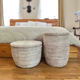 Two white woven baskets with flat lids, one round and short, the other tall and cylindrical, placed side by side on a wooden floor near a bed with neutral bedding.