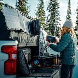 Person in green jacket and gray beanie connecting orange and black Poseidon XL Power Bank to solar panel mounted on white truck's open tailgate in snowy forest.