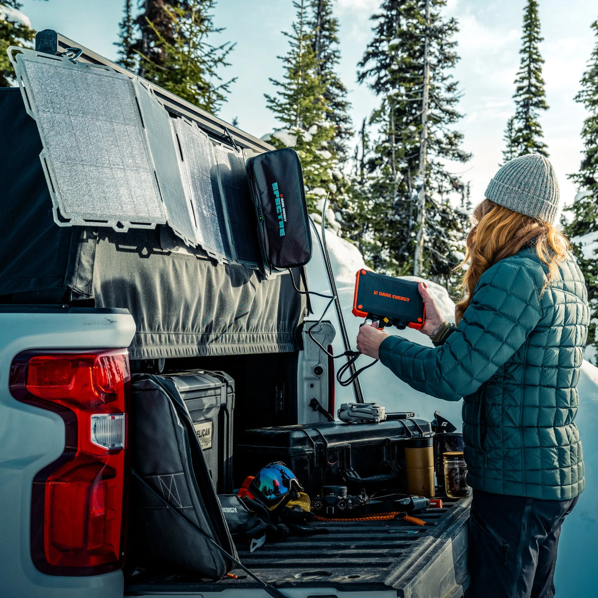 Person in green jacket and gray beanie connecting orange and black Poseidon XL Power Bank to solar panel mounted on white truck's open tailgate in snowy forest.