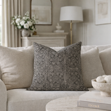 Square pillow with a detailed black and beige floral pattern placed on a light beige fabric sofa in a softly lit living room.
