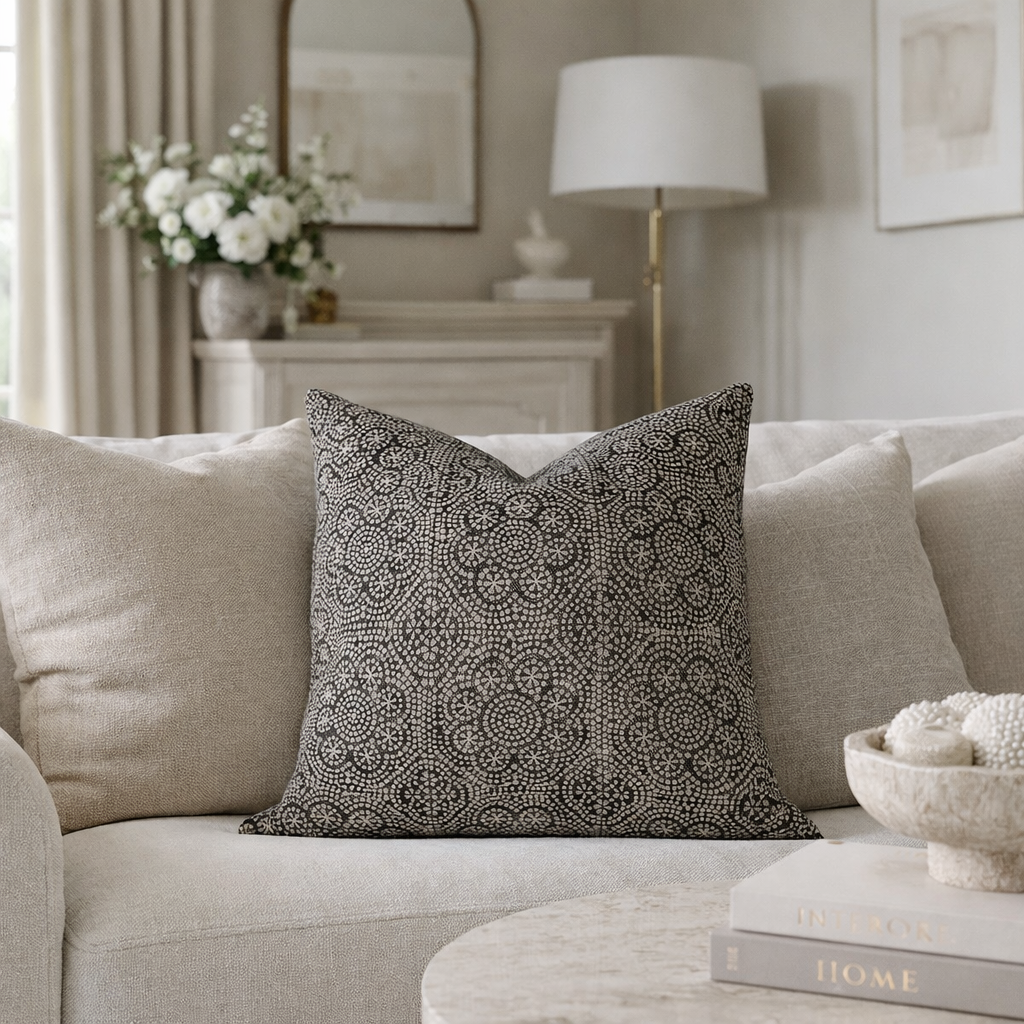 Square pillow with a detailed black and beige floral pattern placed on a light beige fabric sofa in a softly lit living room.
