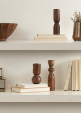 Three wooden taper candle holders with smooth curves and varying heights displayed on white shelves among neutral-toned books and decorative objects.