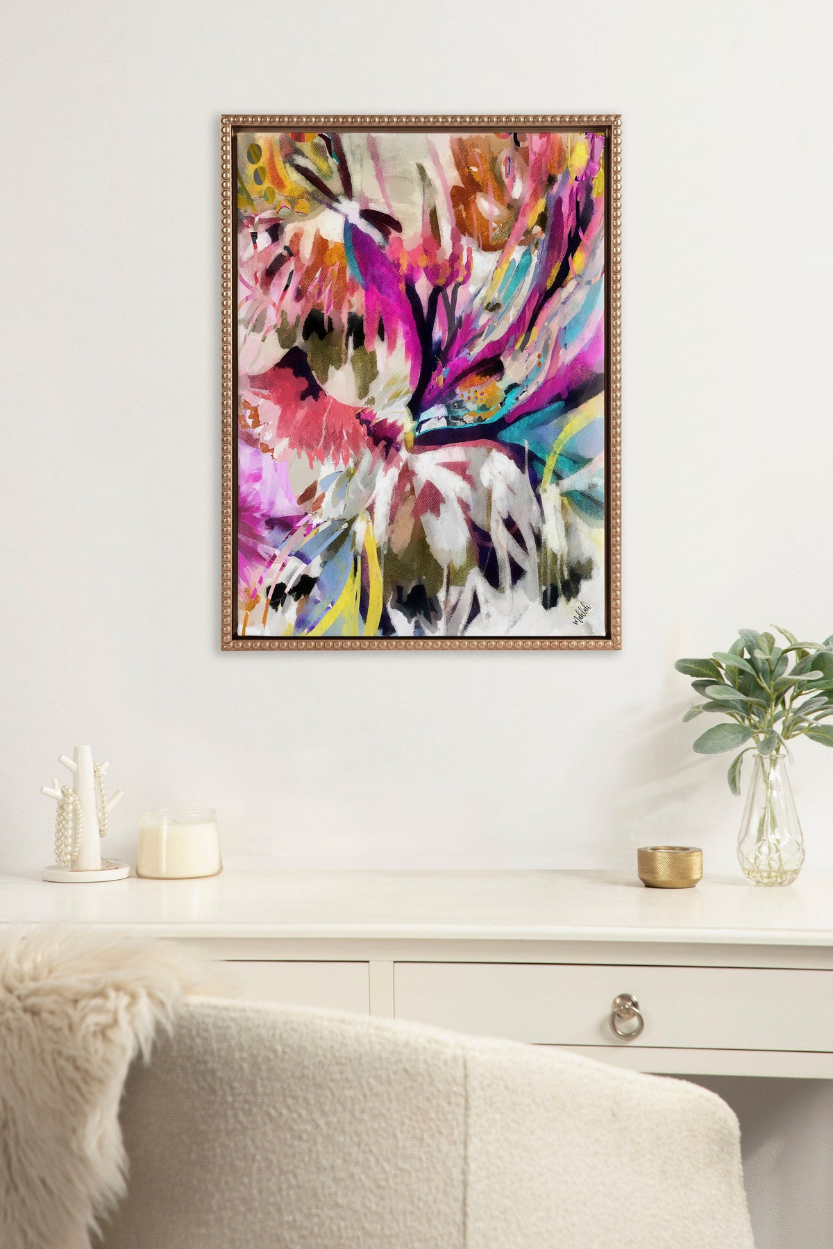 Abstract floral painting with vivid pinks, purples, yellows, and blues in a beaded wooden frame, hanging above a white desk with decorative items and a plant.