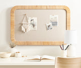 Rectangular beige fabric pinboard with rounded rattan frame hanging above a white desk with open book, white textured lamp, and small vases.