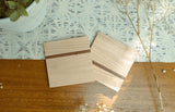 Two square wooden coasters with a single dark wood stripe running through center, shown on a patterned fabric and wooden surface with plant and dried flower accents.