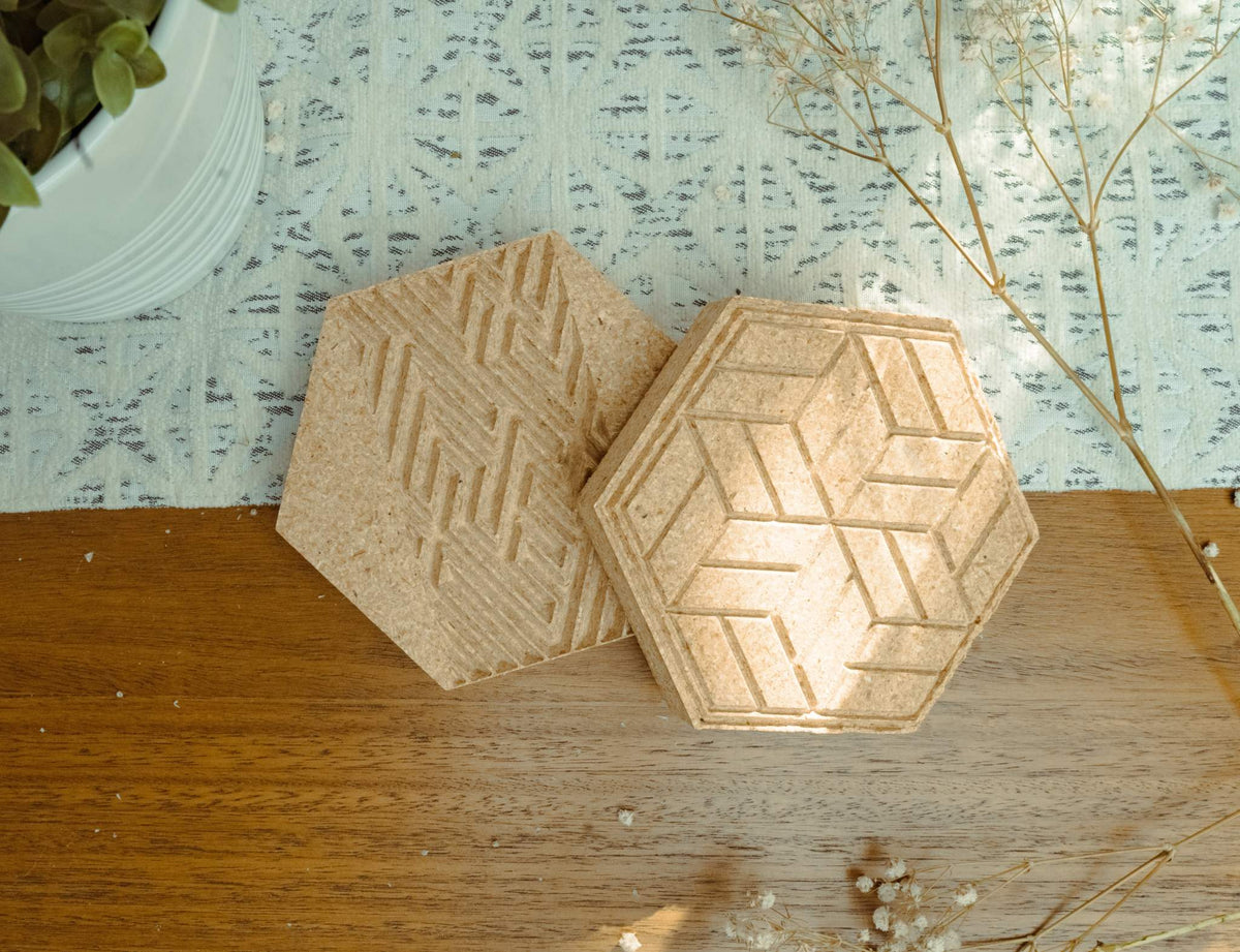Two hexagonal cork coasters with geometric carved patterns, one featuring zigzag lines and the other repetitive hexagonal blocks, on a wooden surface with soft fabric and dried flowers nearby.