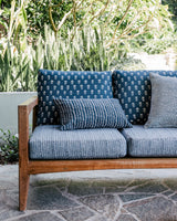 Outdoor wooden sofa with blue seat cushions featuring subtle white stripes and patterned back cushions in darker blue with small white motifs, set against green plants.