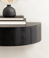 Close-up of a round, black floating wood side table mounted on a white wall, with a matte black vase holding dried flowers and two stacked books on top.