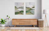 Pair of abstract landscape paintings in soft muted greens, grays, and beiges framed and hung side by side above a light wood sideboard in a minimalist living room.