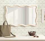 Rectangular wall mirror with a wavy scalloped rattan frame in natural beige, reflecting window panes, above a white console holding a taupe tote, dark wooden bowl, and sunglasses.