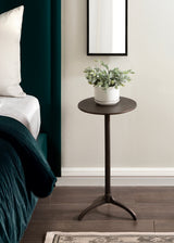 Round dark metal side table with three curved legs holding a white pot with green eucalyptus branches beside a bed with teal bedding and white pillow.
