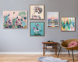 Five colorful framed artworks on a light gray wall above a small wooden table and a modern chair with a pink cushion, showcasing floral, abstract, and sunset themes.