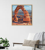 Square framed canvas painting of a red sandstone arch set against blue sky and distant mountains, hanging above a white armchair with neutral pillows and grey throw