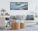 Light neutral-toned living room with beige sofa adorned with gray and blue cushions, natural wood round coffee tables, and a large framed seascape painting overhead.