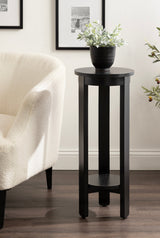 Tall black round wood side table with three legs and a lower circular shelf, topped with a small black planter holding green leafy stems, next to a white upholstered chair.