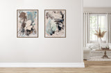 Two abstract paintings with blues, browns, and whites in wooden frames hanging on a white wall, adjacent to a bright living room with a beige sofa and neutral decor.