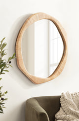 Irregular round wall mirror with a textured natural rattan frame reflecting a sunlit window, next to green foliage and a cozy olive green chair draped with a chunky knit blanket.