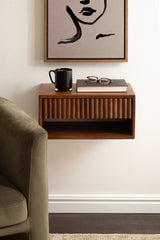 Floating wooden side table with ribbed front panel and open storage below, holding a black mug, closed book, and glasses, mounted on a white wall