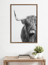 Black and white framed portrait of a Highland cow facing forward with shaggy hair partially covering its eyes and one prominent curved horn on a white wall above a wooden table.
