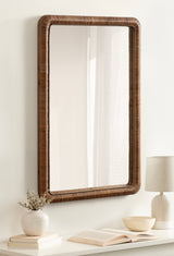 Rectangular wall mirror with a rounded, beaded dark brown rattan frame hanging above a white shelf holding a white vase with dried flowers, an open book, a bowl, and a lamp.