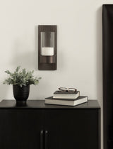 Vertical dark metal wall sconce with ribbed texture holding a glass candle holder, mounted above a black cabinet with books, glasses, and a black pot with greenery