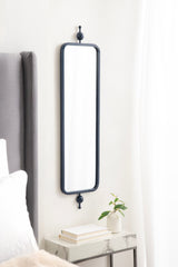 Tall, narrow rectangular mirror with rounded edges and a slender black metal frame, mounted vertically on a light wall between a gray bed and white curtains.