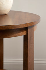 Close-up of a round wood side table's smooth brown tabletop and sturdy rectangular leg against a neutral wall background