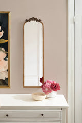 Tall, narrow arched mirror with ornate bronze detailing on top, mounted on a light beige wall above a white dresser holding a small bowl and a white vase with pink flowers.