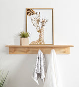 Framed portrait of a mother giraffe nuzzling a baby giraffe with light brown spots on white, hanging above a wooden coat rack with two hanging towels and a small potted plant.