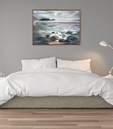 Framed canvas painting of a moonlit seascape with rocks and gentle waves beneath a cloudy sky, displayed above a bed with white bedding and neutral headboard.