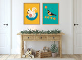 Two colorful framed canvases on a white wall, one with a white squirrel on mustard yellow, the other with a black bird on teal, above a rustic wooden console table.