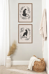 Two vertically stacked framed canvases featuring black spotted jungle cats surrounded by simple black leaf designs on a neutral linen background, arranged on a pale wall.
