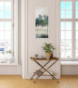 Tall framed canvas print of abstract trees in blue, green, and gray tones hanging above a gold and wood bar cart holding a small potted plant and decorative items in a bright room.