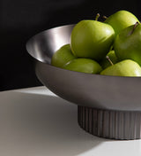 Matte silver metal bowl with vertical ribbing on the short cylindrical base, holding bright green apples, set against a black background and white surface.