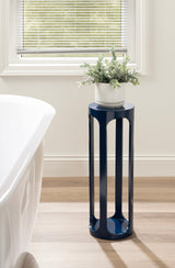 Tall, cylindrical navy blue stool with four vertical open panels, topped with a white pot holding green leafy plants, standing on light wood flooring near a window and bathtub.