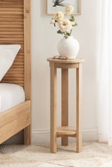 Tall, round wooden side table with three legs and a lower circular shelf, holding a white vase with cream flowers and glasses, next to a wooden bed frame.