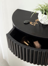 Close-up of black semi-circular side table with ribbed front edge, showing open drawer containing makeup items and a white geometric planter with green plant on top.